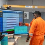 A person in orange coveralls using a large digital display in a workshop.