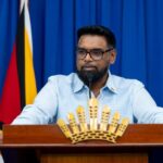 President Irfaan Ali speaking at a lectern, with a blue curtain and Guyana flag behind him.