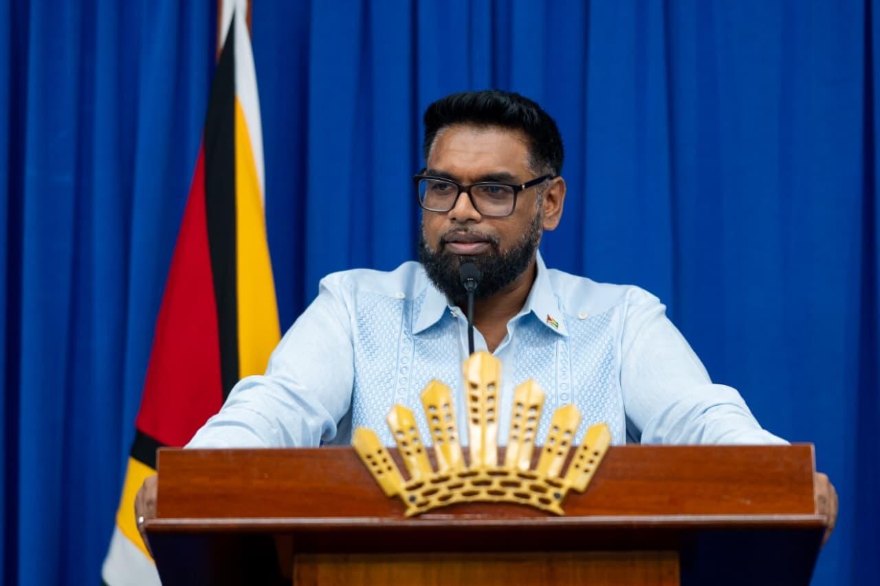 President Irfaan Ali speaking at a lectern, with a blue curtain and Guyana flag behind him.